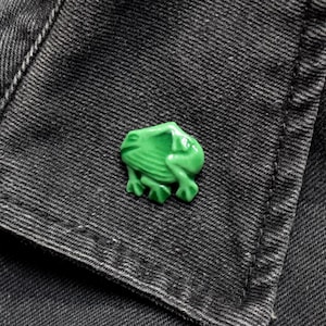 Puede incluir: Un broche en forma de rana de color verde brillante sujeto a una tela de mezclilla gris oscuro. El broche presenta un acabado liso y brillante con detalles moldeados que representan la cabeza, el cuerpo y las patas de la rana en cuclillas.