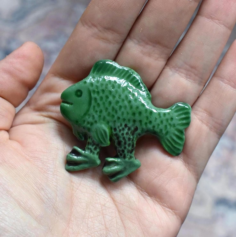 Puede incluir: Una peque&ntilde;a figura de cer&aacute;mica verde brillante con forma de pez y dos pies palmeados. La superficie presenta un patr&oacute;n texturizado y con hoyuelos en el cuerpo y las aletas, con un acabado de esmalte liso y brillante que refleja la luz.