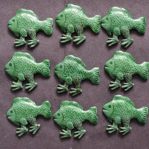 Puede incluir: Un conjunto de nueve botones de cer&aacute;mica verde con forma de pez, dispuestos en una cuadr&iacute;cula de tres por tres. Cada pez presenta un acabado brillante, un patr&oacute;n de escamas texturizado en el cuerpo y dos patas palmeadas distintas.