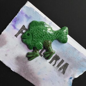 Puede incluir: Una figura de pez de cer&aacute;mica verde brillante con escamas texturizadas y dos pies peque&ntilde;os, colocada sobre un papel rectangular con un lavado de acuarela en tonos azules y morados, con el texto impreso en negro parcialmente visible FLORIA.