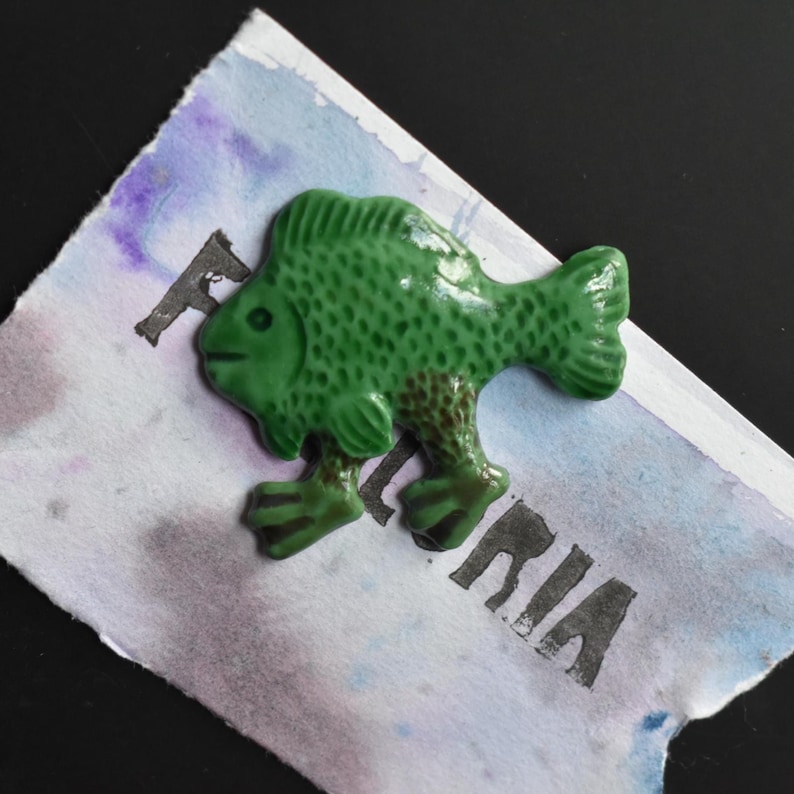 Puede incluir: Una figura de pez de cer&aacute;mica verde brillante con escamas texturizadas y dos pies peque&ntilde;os, colocada sobre un papel rectangular con un lavado de acuarela en tonos azules y morados, con el texto impreso en negro parcialmente visible FLORIA.