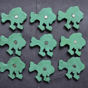 Puede incluir: Un conjunto de nueve imanes con forma de pez dispuestos en una cuadr&iacute;cula de tres por tres. Cada figura de pez presenta una superficie verde mate y lisa con un peque&ntilde;o im&aacute;n circular de color plateado incrustado en el centro del cuerpo.