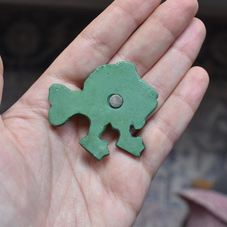 Puede incluir: Un peque&ntilde;o objeto verde texturizado con forma de pez que tiene un im&aacute;n circular de color plateado incrustado en el centro. La superficie del material verde presenta un patr&oacute;n de puntos fino y uniforme en toda su forma, incluyendo aletas y cola.