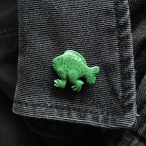May include: A small, bright green, three-dimensional fish-shaped button with visible fins and legs, featuring a textured surface with small indentations. The button is attached to the dark grey, ribbed fabric of a garment cuff.