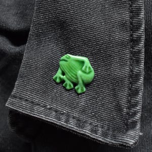 May include: A bright green frog-shaped button with a smooth, glossy finish and moulded details depicting the frog's limbs and body. The button is attached to a piece of dark grey, textured denim fabric with visible stitching along the edge.