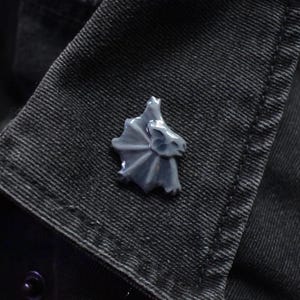 Puede incluir: Un pequeño pin de esmalte gris claro con forma de lagarto de cuello con volantes, con un volante prominente en el cuello y un perfil de cabeza detallado. El pin está sujeto al cuello de tela texturizada gris oscuro de una prenda, mostrando un acabado liso y brillante.