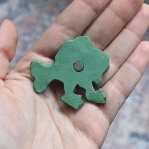 Puede incluir: Un objeto verde texturizado con forma de pez que tiene un peque&ntilde;o im&aacute;n met&aacute;lico circular incrustado en el centro. La superficie del objeto presenta un patr&oacute;n de l&iacute;neas cruzadas finas y la forma general muestra una estructura clara de cola, cuerpo y aleta.