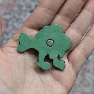 Puede incluir: Un im&aacute;n verde mate con forma de pez y superficie texturizada. El im&aacute;n presenta un peque&ntilde;o disco met&aacute;lico circular de color plateado incrustado en el centro del cuerpo del pez, que tiene una aleta caudal distintiva y dos aletas inferiores.