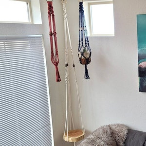 May include: Three macrame plant hangers in burgundy, white, and navy blue. The burgundy hanger is empty, the white one has a wooden seat, and the navy blue one holds a small potted plant. All are suspended from wooden rings.