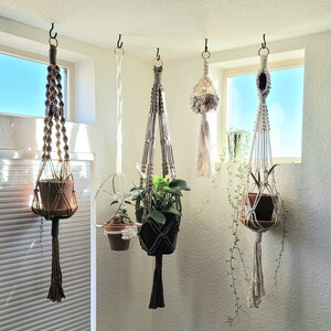 May include: Five macrame plant hangers of varying designs and colors are suspended from black hooks near a window. The hangers hold potted plants, showcasing a bohemian aesthetic. The hangers feature intricate knotting and tassel details.