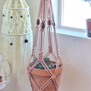 May include: A handmade macrame plant hanger in a soft pink hue, adorned with dark wooden beads. The plant holder suspends a terracotta pot filled with small succulents and cacti. A second, off-white macrame piece hangs in the background.