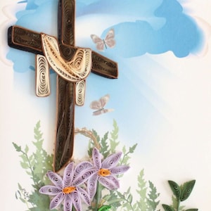 May include: A paper quilling artwork featuring a brown cross with a cream-colored drape, butterflies, and purple flowers with orange centers. The background includes a blue sky and green foliage.