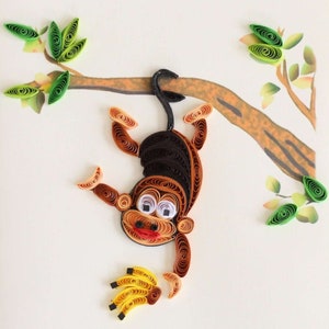 May include: A whimsical quilled paper art piece featuring a brown monkey hanging from a tree branch. The monkey has a smiling face, holding a bunch of yellow bananas. The artwork includes green leaves and is set against a white background.