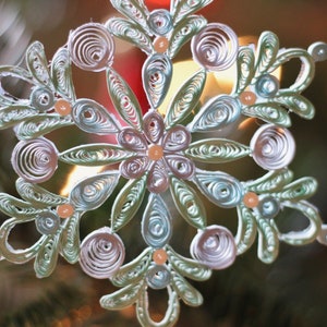 May include: A delicate, white and green paper snowflake ornament with pearl-like beads. The snowflake is intricately designed with multiple layers of paper, creating a 3D effect.