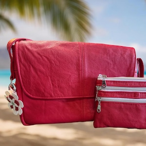 May include: A red leather shoulder bag and matching small zippered pouch. The bag has a flap closure and a decorative flower charm. The pouch has three silver zippers. The background shows a beach scene with palm leaves and the text "UpBeat Bags".