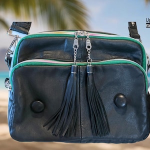 Puede incluir: Bolso bandolera de cuero negro con ribete verde y rosa, cremalleras plateadas y dos borlas negras. El bolso tiene una correa para el hombro y dos botones negros redondos. El texto "UpBeat Bags" y "Kāneʻohe, Hawaiʻi USA" son visibles.