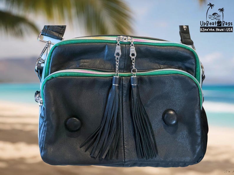 Puede incluir: Bolso bandolera de cuero negro con ribete verde y rosa, cremalleras plateadas y dos borlas negras. El bolso tiene una correa para el hombro y dos botones negros redondos. El texto "UpBeat Bags" y "Kāneʻohe, Hawaiʻi USA" son visibles.