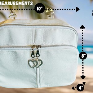 May include: White leather bag with silver zippers and heart-shaped zipper pulls. The bag is 10 inches wide, 8 inches tall, and 4 inches deep. The bag has a top handle and a front pocket. The background shows a beach scene.