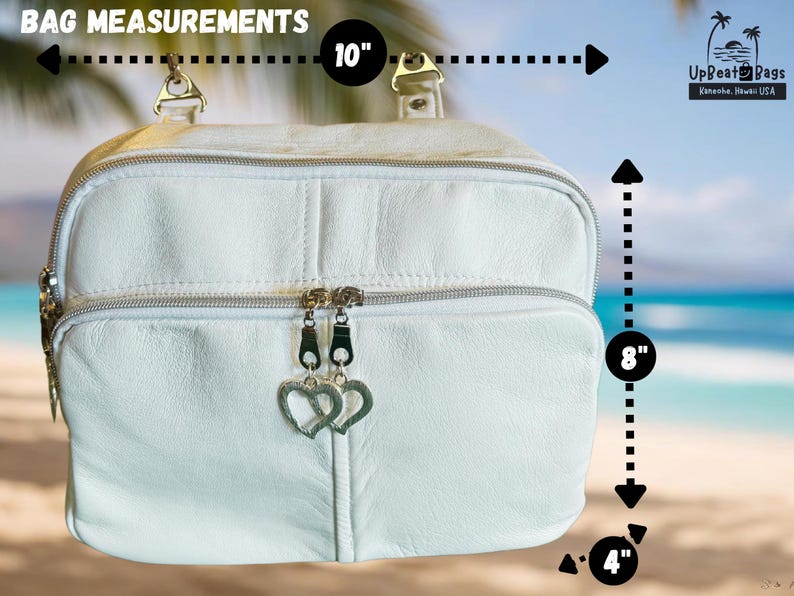 May include: White leather bag with silver zippers and heart-shaped zipper pulls. The bag is 10 inches wide, 8 inches tall, and 4 inches deep. The bag has a top handle and a front pocket. The background shows a beach scene.