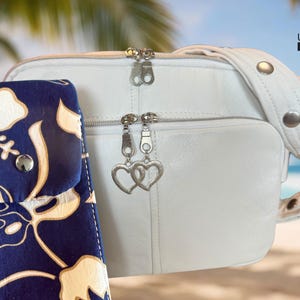 May include: A white leather crossbody bag with a matching strap and silver hardware. A navy blue and white floral print case is next to the bag. The bag has two zipper compartments and heart-shaped charms.