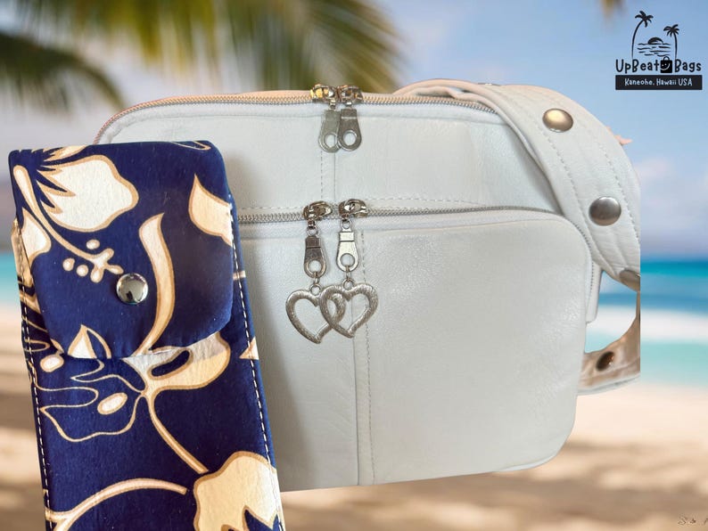 May include: A white leather crossbody bag with a matching strap and silver hardware. A navy blue and white floral print case is next to the bag. The bag has two zipper compartments and heart-shaped charms.