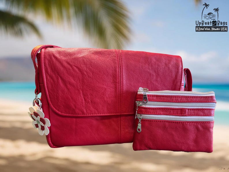 May include: A red leather shoulder bag and matching small zippered pouch. The bag has a flap closure and a decorative flower charm. The pouch has three silver zippers. The background shows a beach scene with palm leaves and the text "UpBeat Bags".