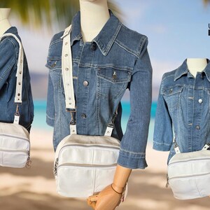 May include: A white leather crossbody bag with a silver zipper and a white adjustable strap. The bag is paired with a denim jacket. The bag has a rectangular shape with a front pocket and a top zipper.