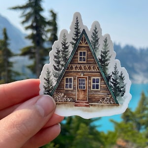 May include: A watercolour illustration of a brown wooden A-frame cabin with a green roof and a white door. The cabin is surrounded by green trees and has a wood-burning stove and a stack of firewood.