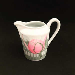 May include: White ceramic creamer with a pink and green floral design. The creamer has a rounded body and a handle.