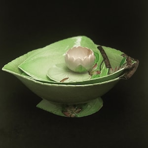 May include: A set of three green ceramic bowls shaped like lily pads, with a small white ceramic lily flower in the centre. The bowls are decorated with green leaves and pink flowers.