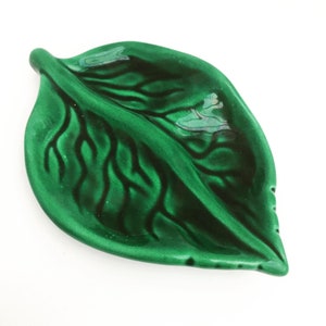 Puede incluir: Un plato de cerámica verde brillante con forma de hoja. El plato presenta vetas detalladas y un color verde intenso y rico. El diseño de la hoja es un acento decorativo para el hogar.