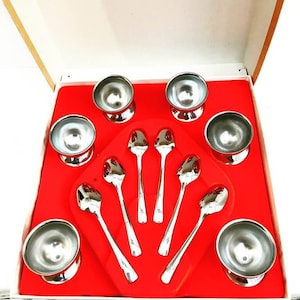 Puede incluir: Un juego de seis cuencos de helado de plata y seis cucharas de plata en una caja roja y blanca. Los cuencos son redondos y tienen una base pequeña. Las cucharas son largas y tienen un extremo redondeado.