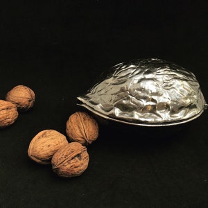 Puede incluir: Un cascanueces de plata con forma de nuez y superficie texturizada. Cuatro nueces están esparcidas sobre una superficie negra.