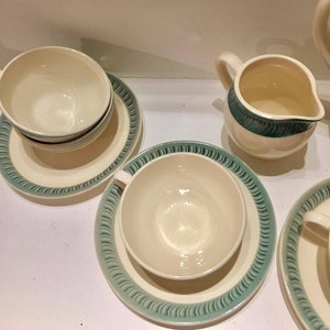 May include: A set of cream-colored ceramic tea cups and saucers with a teal-colored rim featuring a repeating diagonal line pattern. A matching cream-colored creamer pitcher with a teal band around the top rim is also included in the set.