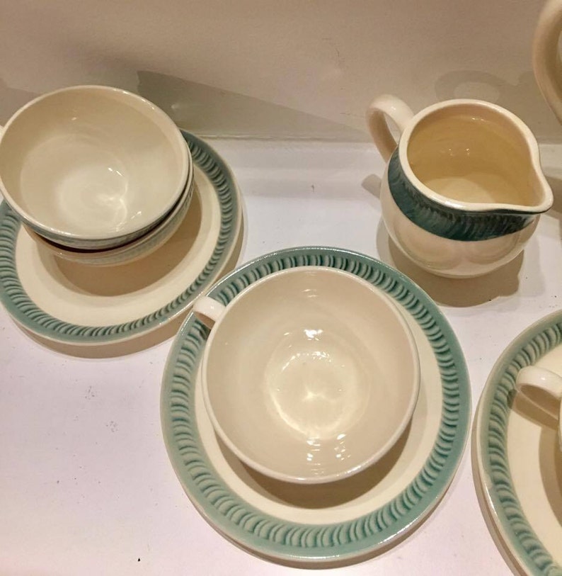 May include: A set of cream-colored ceramic tea cups and saucers with a teal-colored rim featuring a repeating diagonal line pattern. A matching cream-colored creamer pitcher with a teal band around the top rim is also included in the set.
