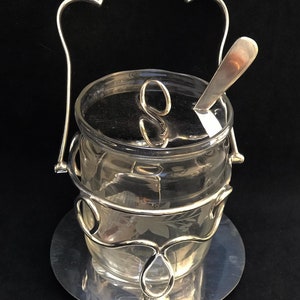 Könnte beinhalten: Eine klare Zuckerdose aus Glas mit einem silberfarbenen Metallgriff, Deckel und Boden. Die Schüssel hat ein floral geätztes Design und einen kleinen Löffel. Der Griff hat eine dekorative, geschwungene Oberseite. Die Schüssel steht auf einem runden Sockel.