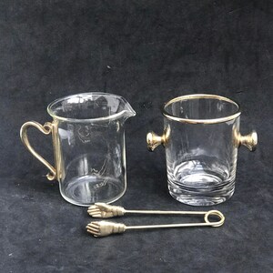 Peut inclure: Un ensemble de trois articles de bar en verre transparent, comprenant un pichet, un seau et une paire de pinces. Le pichet et le seau ont des bords dorés. Les pinces ont des poignées dorées en forme de mains.