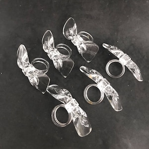 Könnte beinhalten: Sechs transparente Serviettenringe aus Kunststoff in Form von Schleifen. Die Ringe sind so konzipiert, dass sie Servietten auf einem Esstisch an Ort und Stelle halten.
