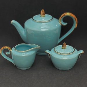 Könnte beinhalten: Ein Teekannen-, Milchkännchen- und Zuckerdose-Set in Tealblau mit goldenen Akzenten. Die Teekanne hat einen geschwungenen Griff und ein goldenes Blattdesign auf dem Deckel. Das Milchkännchen und die Zuckerdose haben goldene Ränder.