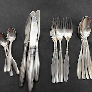 Può includere: Una collezione di posate in acciaio inossidabile, tra cui cucchiai, forchette e coltelli, disposte su una superficie scura. Le posate hanno un design semplice ed elegante con una serie di linee orizzontali vicino alla base dei manici.