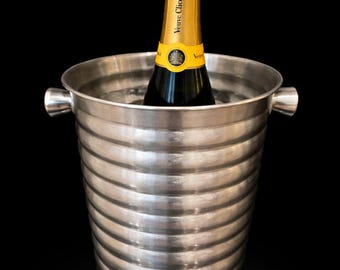 May include: A silver champagne bucket with a ridged design holds a bottle of Veuve Clicquot champagne. The bottle has a gold foil top and a yellow label. The bucket has two small handles and sits against a black background.