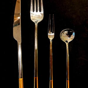 May include: A collection of four silver-coloured eating utensils: a knife, a large fork, a small fork, and a soup spoon. Each piece has a gold-coloured accent at the base of the handle. The background is black.