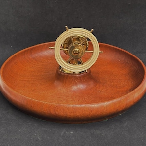 May include: A wooden bowl with a brass ship's wheel nutcracker in the center. The bowl is a reddish brown color and the wheel is gold.