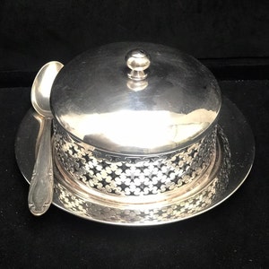 Puede incluir: Un mantequillero de plata con tapa abovedada y una cucharita. El plato tiene un diseño decorativo calado alrededor de la base.