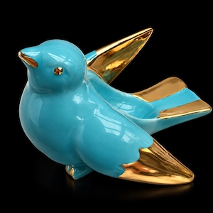 Peut inclure: Une figurine d'oiseau en céramique avec une finition turquoise brillante et des accents en or métallique sur le bout des ailes, les plumes de la queue et le bec. L'oiseau est représenté en position perchée, ailes légèrement déployées.