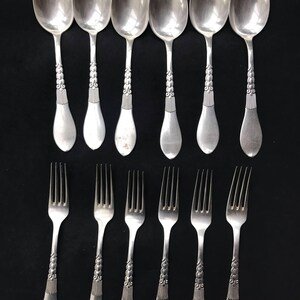 May include: A set of vintage silverware, including six spoons and six forks, arranged on a black surface. The spoons and forks are silver-colored with ornate, detailed handles. The spoons have a rounded bowl shape, and the forks have four tines.