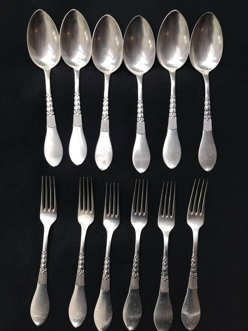 May include: A set of vintage silverware, including six spoons and six forks, arranged on a black surface. The spoons and forks are silver-colored with ornate, detailed handles. The spoons have a rounded bowl shape, and the forks have four tines.