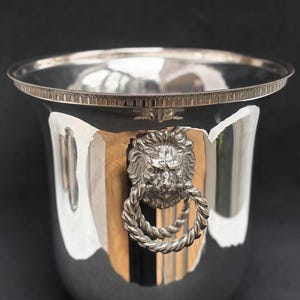 Puede incluir: Cubeta de vino plateada con asa de cabeza de león. La cubeta tiene un borde ancho y acanalado y una superficie pulida que refleja el entorno. La cabeza de león es detallada, con un asa de cuerda trenzada.