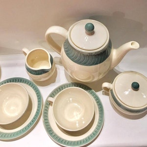 May include: A ceramic tea set featuring a teapot with a lid, a creamer, a sugar bowl with a lid, and two teacups with matching saucers. Each piece is cream-colored with a teal-colored band decorated with a repeating, textured, diagonal brushstroke pattern.