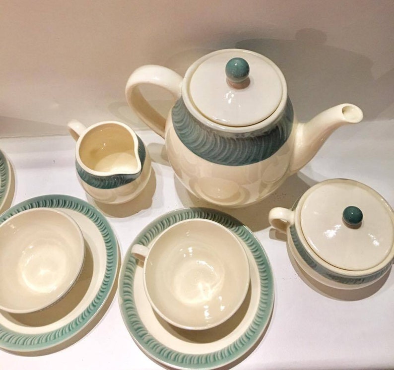 May include: A ceramic tea set featuring a teapot with a lid, a creamer, a sugar bowl with a lid, and two teacups with matching saucers. Each piece is cream-colored with a teal-colored band decorated with a repeating, textured, diagonal brushstroke pattern.